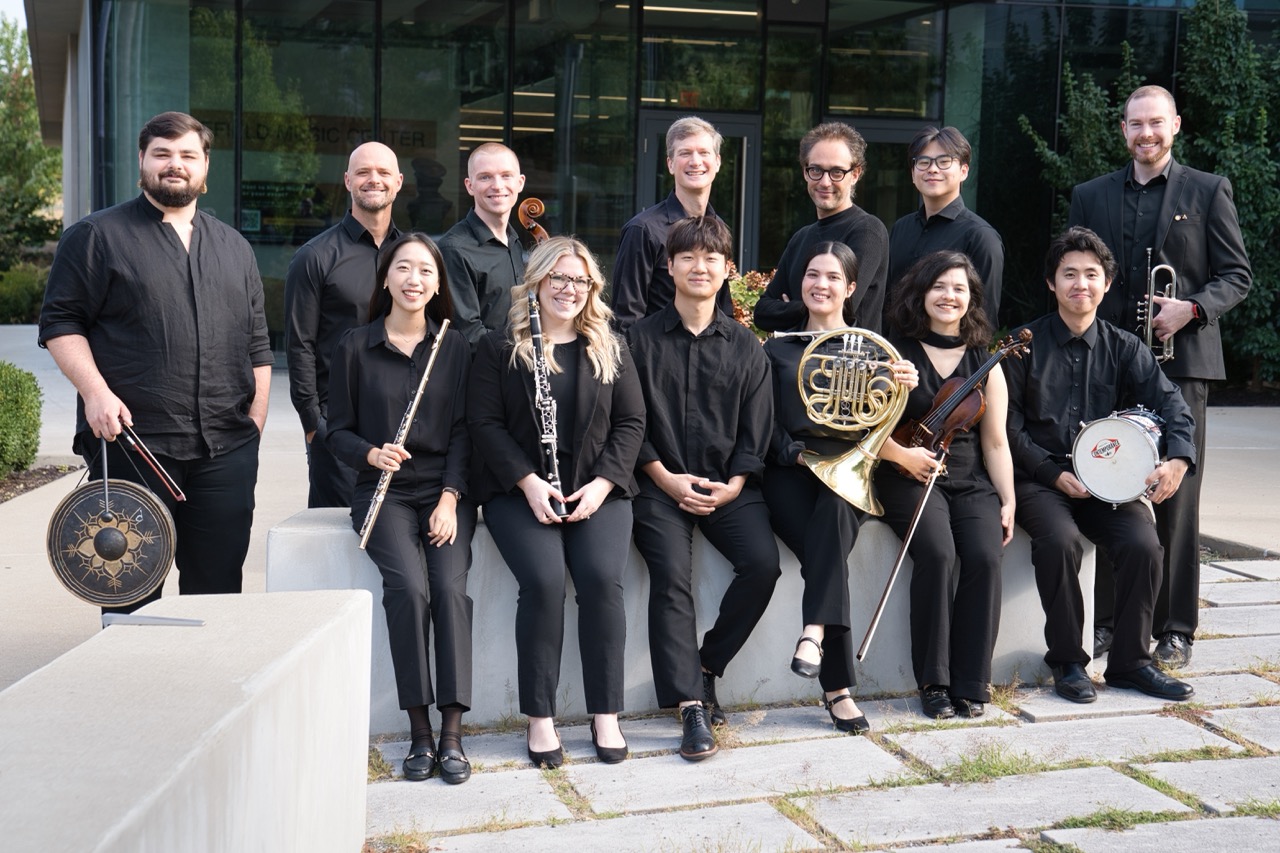 A group of eleven musicians poses outdoors, smiling in black attire. They hold various instruments, conveying a sense of unity and professionalism.