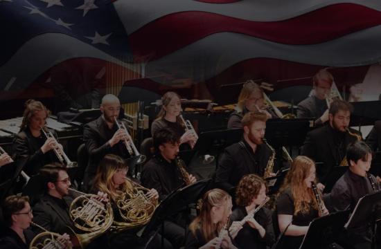 Concert band musicians in black perform on brass and woodwind instruments beneath a large American flag.